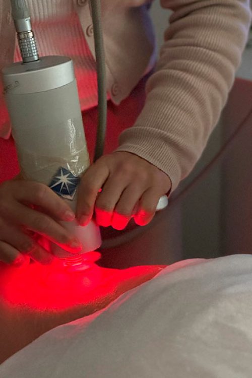 Una persona tumbada en una camilla y otra aplicándole un aparato con luz roja sobre la cara